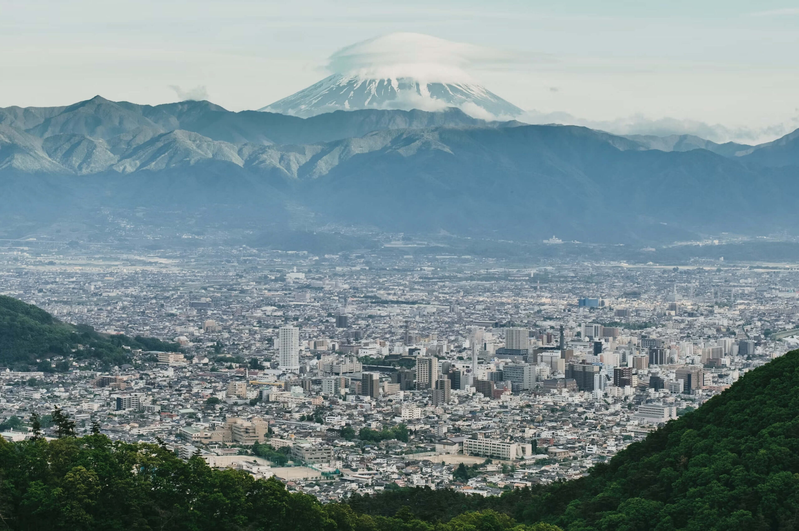 甲府盆地を見下ろす高台から、眼下に広がる市街地と、遠方に連なる山々の背後にそびえる冠雪の富士山。
