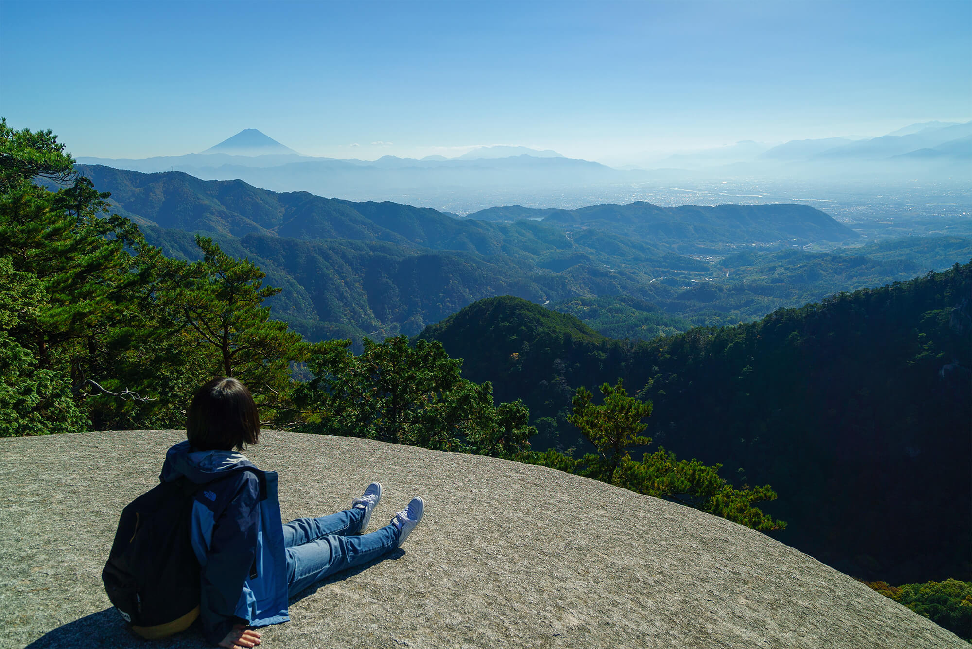 弥三郎岳の大きな岩場に腰掛け、遠くの山並みと富士山を眺めるリュックを背負った人物